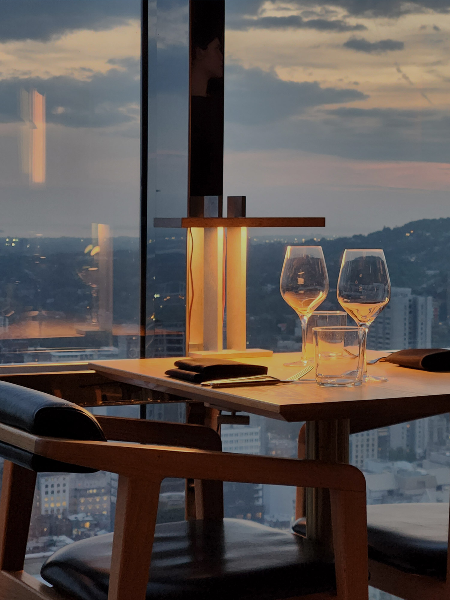 Dining-T Table set for two in a high-rise restaurant with two wine glasses, warm lamp, and city skyline at dusk outside the window