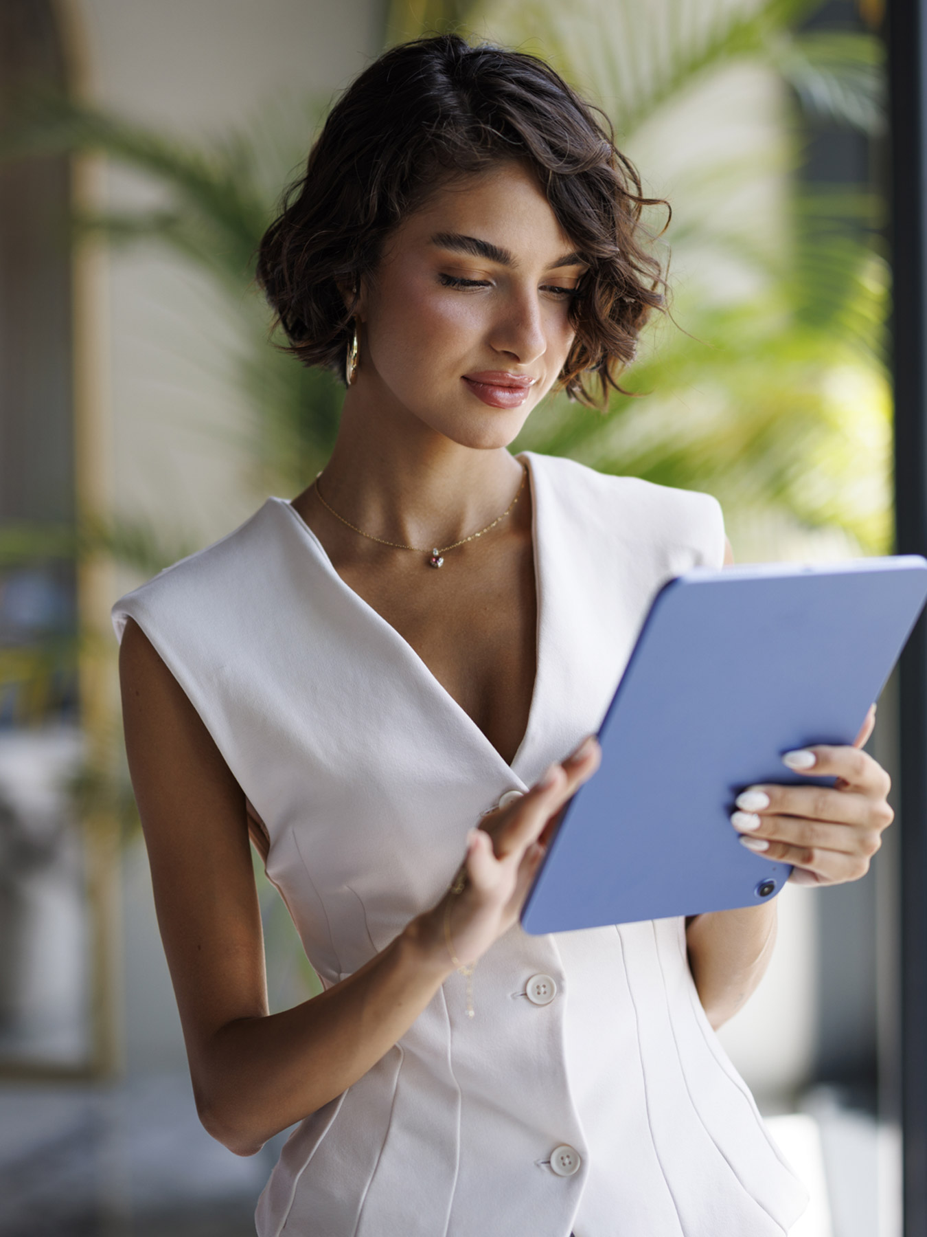 FOH-T Woman in a white sleeveless dress holding a blue tablet, looking at it outdoors by greenery.