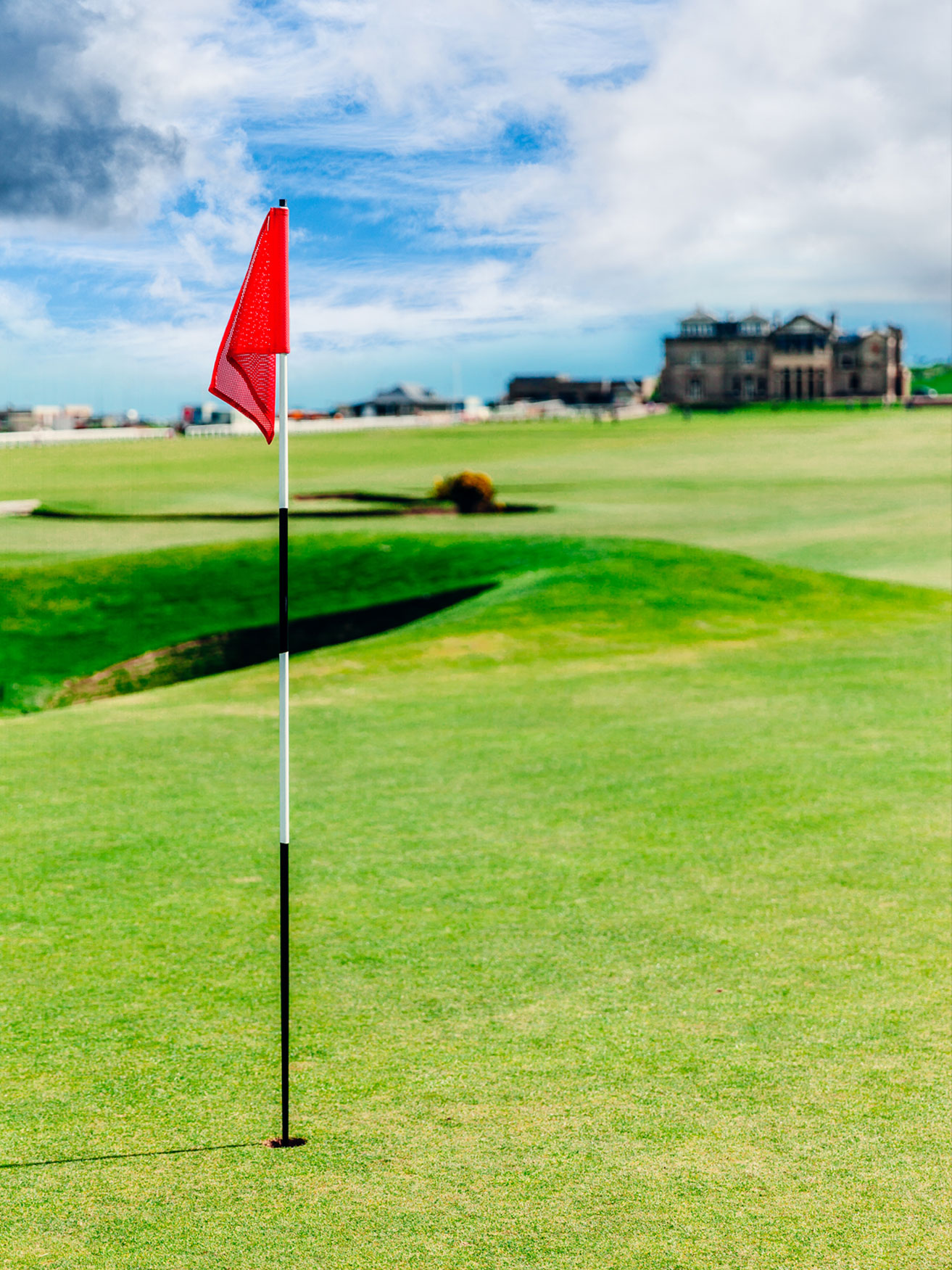 Golf-Flag Red flag on a white and black golf flagstick marking a hole on a green, with a distant building in the background.