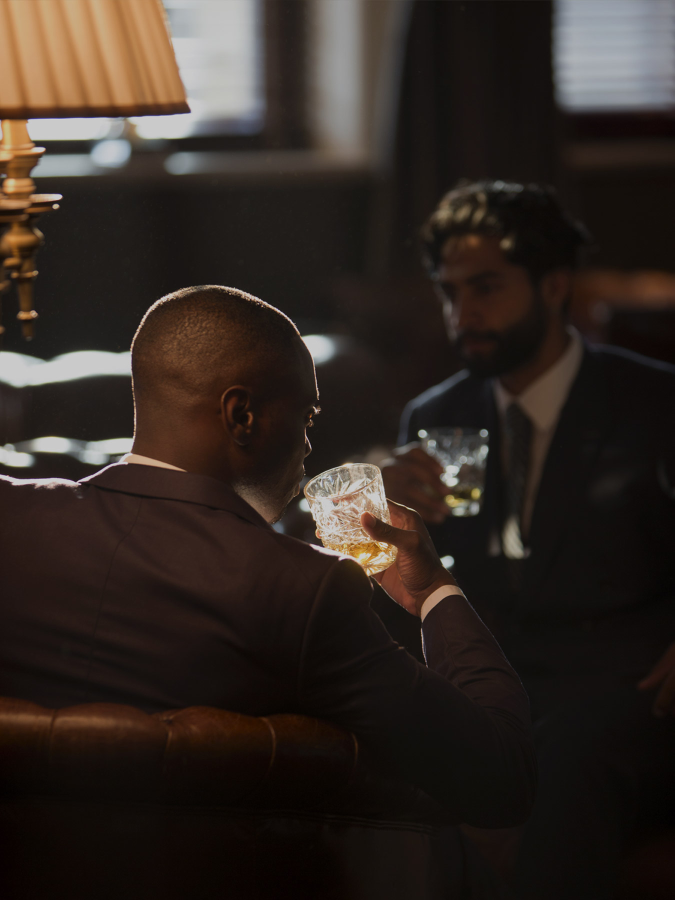 Private-Members-T Man in a suit toasts with a crystal glass in a dim lounge, another man blurred in the background.